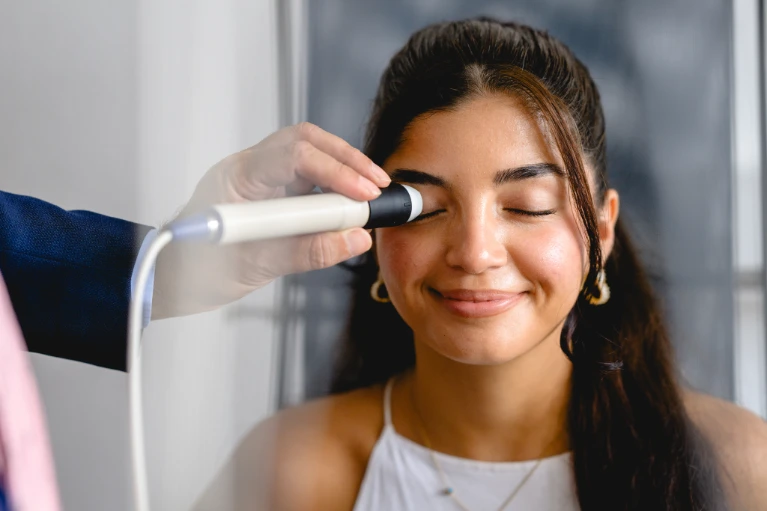 Eine Frau mit langem dunklem Haar lächelt mit geschlossenen Augen, während ein Gerät an ihr rechtes Auge gehalten wird.