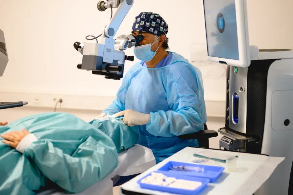 Chirurg in blauer OP-Kleidung führt Augenoperation mit Mikroskop durch; Patient unter grünem Tuch, Instrumente und Monitore sichtbar.