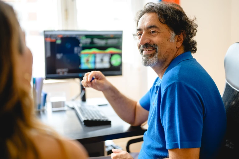 Mann im blauen Polo am Schreibtisch, lächelnd, gestikuliert vor Computer mit Grafiken gegenüber einer Frau im Büro.