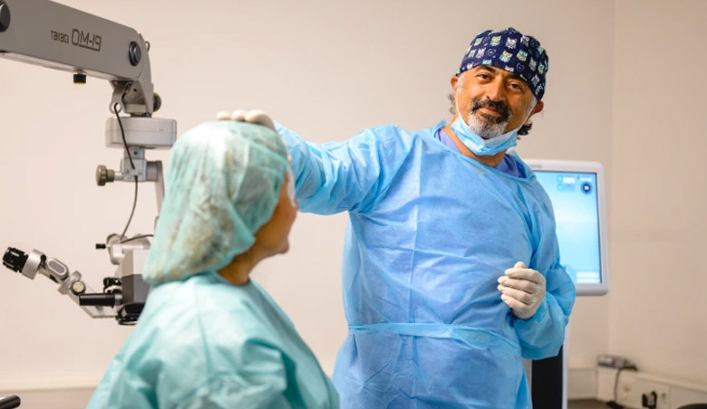 Ein Chirurg in blauem Kittel berührt sanft den Kopf eines sitzenden Patienten in einem Untersuchungsraum mit medizinischen Geräten.