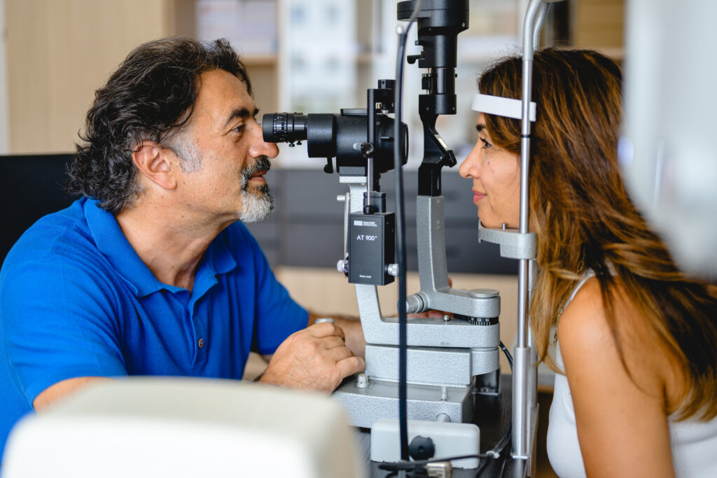 Ein Mann im blauen Hemd untersucht mit einer Spaltlampe die Augen einer Frau in einer Klinik; sie stützt ihr Kinn auf das Gerät, um sich untersuchen zu lassen.