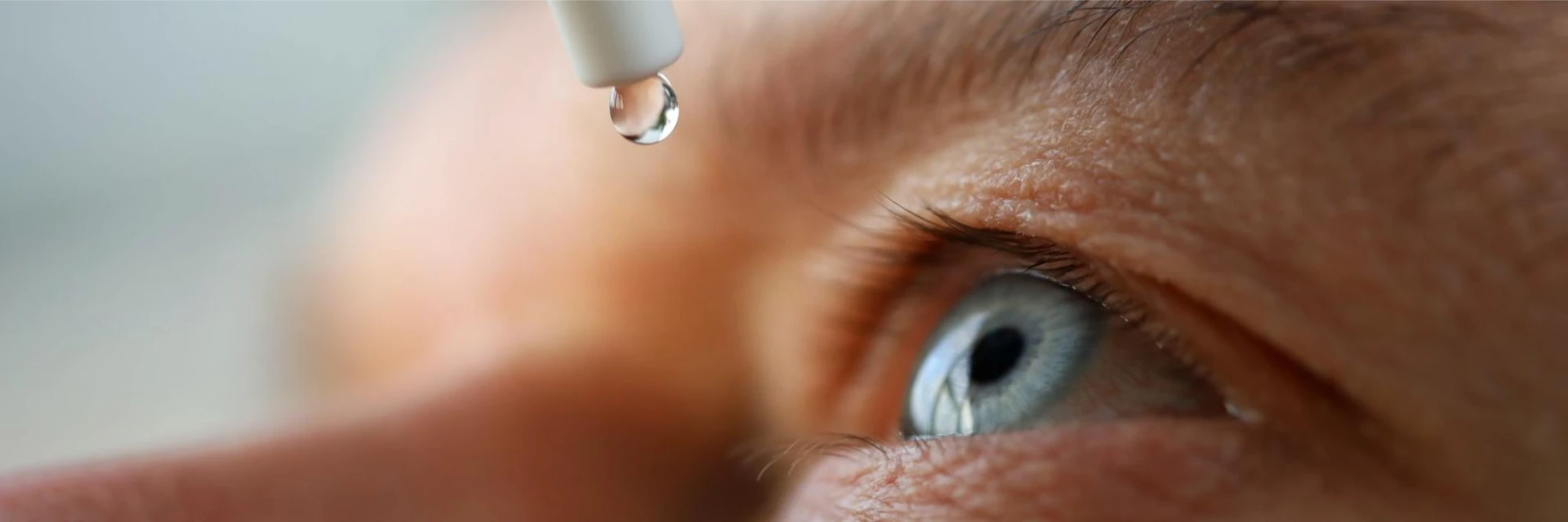 Eine Pipette schwebt über dem blauen Auge einer Person, die gerade einen Tropfen abgibt.