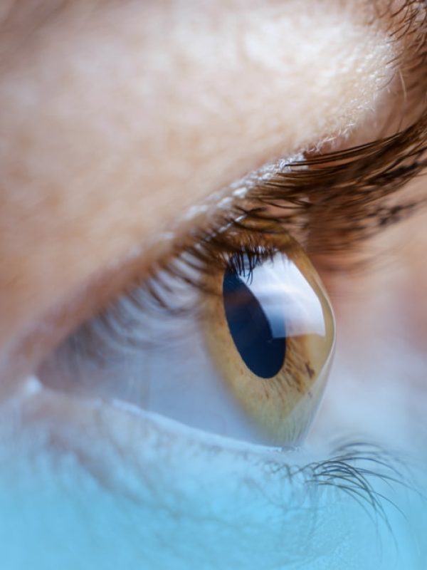 Nahaufnahme eines linken Auges mit hellbrauner Iris, sichtbaren Details und Reflexionen, deutlichen Wimpern und natürlichem Hautton.